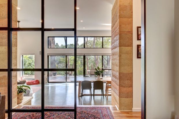 View through a tall black-framed glazed partition or doorway into an open-plan dining and living area. The space features polished concrete flooring, warm timber flooring at the threshold, a large patterned rug, extensive windows overlooking trees, recessed ceiling lighting, a timber dining table with dark chairs, and a prominent exposed horizontal-banded stone or masonry feature wall. The bright interior has a warm, contemporary character.