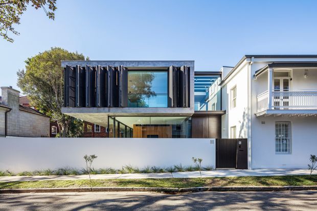 Street-facing view of a contemporary two-storey residence inserted beside a traditional white neighbouring house. The main home has a projecting upper-level rectangular volume with dark vertical louvres, a large glazed opening, exposed concrete-toned framing, and a glazed lower level behind a white masonry boundary wall. A narrow blue-toned glazed link or roof element connects toward the adjoining structure. The scene is bright and urban residential, with restrained modern detailing contrasting against the ornate traditional facade next door.