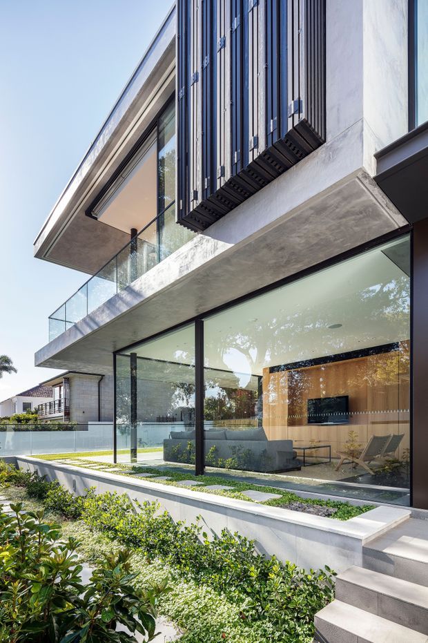 Modern multi-level residential exterior with extensive floor-to-ceiling glazing, projecting concrete slabs, dark vertical metal sun-shading fins, and a glass balustrade. The transparent ground floor reveals a refined living room with built-in cabinetry and a television. Landscaped planting borders the stepped entry and terrace, with a small pool or water feature visible beside the house.
