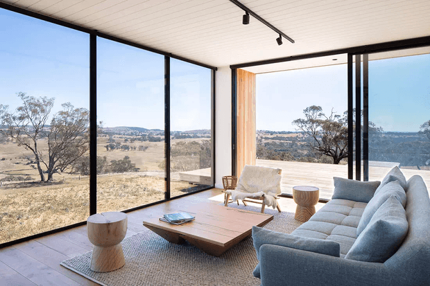 A bright, contemporary living room with floor-to-ceiling black-framed glazing overlooking a dry rural landscape. The room features a large pale sectional sofa, timber coffee table, woven rug, sculptural side tables, a light upholstered lounge chair, white slatted ceiling, and minimalist black track lighting. Large sliding glass openings connect the living area to an outdoor deck.