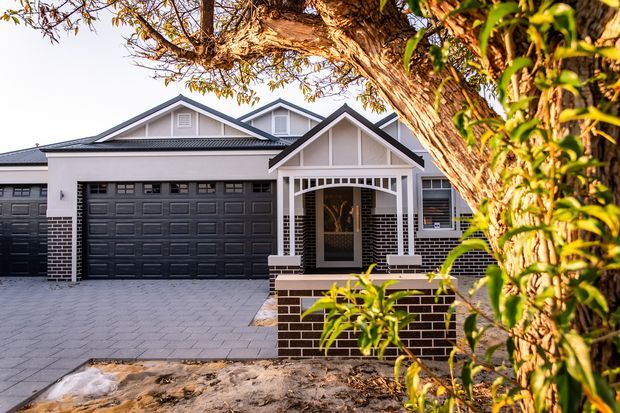 Front exterior view of a contemporary suburban single-storey home with a prominent gabled entry porch, white trim, dark brickwork, multiple garage doors, and a broad paved driveway. Mature trees partially frame the facade.
