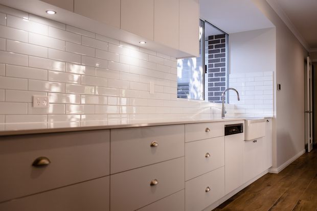 A compact, contemporary kitchen with a long run of pale flat-panel cabinetry, a white glossy subway-tile splashback, integrated appliances, and a stainless-steel gooseneck tap. Recessed ceiling lights illuminate the cabinetry and timber-look flooring, while a window provides natural light and a view of brickwork outside.