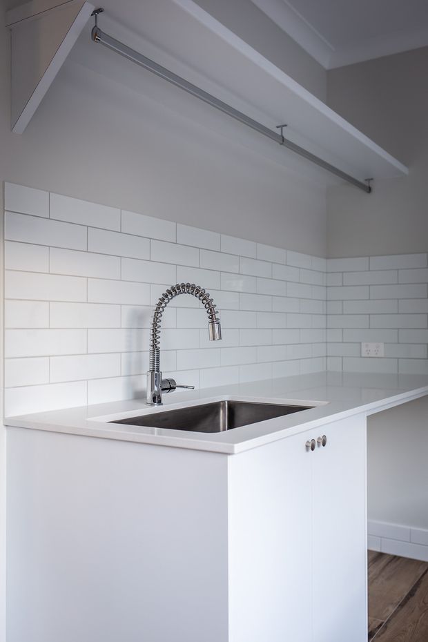 Bright, compact modern kitchen featuring a white island with an integrated stainless-steel double-bowl sink, spring-coil chrome tap, white subway-tile splashback, white cabinetry, and a pale engineered-looking benchtop. A ceiling-mounted rail light and exposed white ceiling beam add functional architectural detail, with timber flooring visible at the edge.