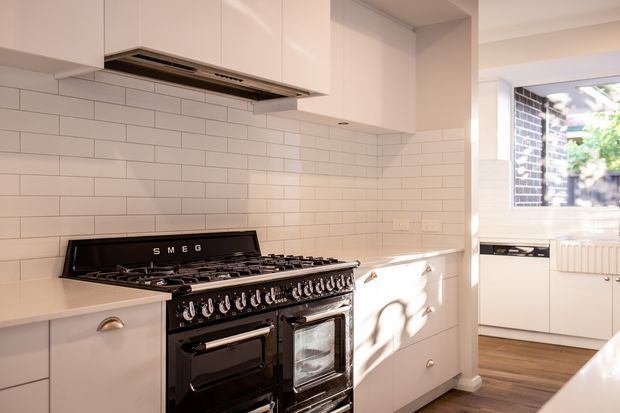 Bright, compact contemporary kitchen featuring white flat-panel cabinetry, a white subway-tile backsplash, pale benchtops, stainless-steel rangehood, and a prominent black Smeg freestanding range cooker. Timber flooring and a windowed utility area are visible beyond the kitchen.
