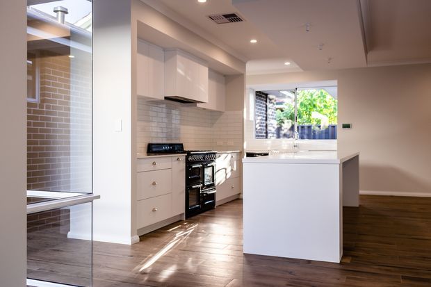 Bright contemporary kitchen viewed from an adjoining open-plan living area. The kitchen features white built-in cabinetry, a tiled splashback, dark freestanding range cooker, central island, timber-look flooring, recessed ceiling lights and a large window overlooking an outdoor area. A glazed opening at left reflects or reveals the kitchen and adjoining spaces.