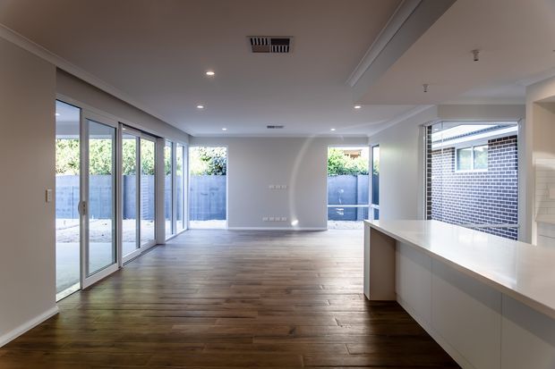 Bright open-plan interior with a spacious living and dining area flowing into a modern kitchen. The room features warm timber flooring, white walls and ceilings with simple cornice detailing, recessed downlights, extensive sliding glass doors along one side, and a clean white kitchen island. A small paved courtyard is visible through the glazing.