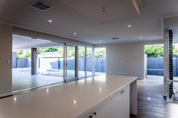 Bright contemporary open-plan kitchen with a large pale island bench and white cabinetry, overlooking a covered outdoor entertaining area and swimming pool through extensive floor-to-ceiling glazing. Dark timber-look flooring, recessed ceiling lighting, and clean white walls create a light, modern interior.