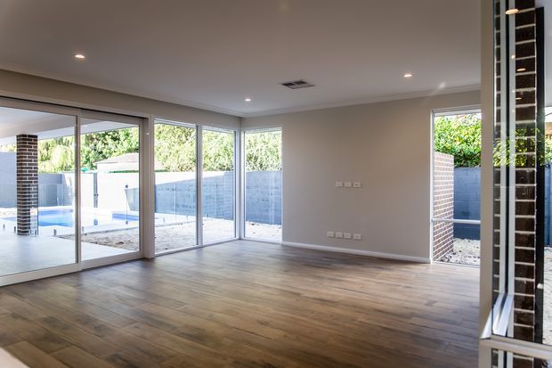 Bright, empty open-plan interior with warm timber flooring, neutral painted walls, recessed ceiling lights, and extensive floor-to-ceiling glazing and sliding doors opening toward a paved outdoor area and swimming pool. Dark brick columns and a partial reflective/glazed feature wall are visible at the right edge.