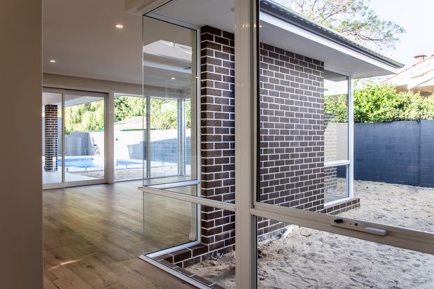 Bright contemporary interior looking through extensive corner glazing and glass doors toward a partially enclosed outdoor courtyard. The space features dark brick walls, warm timber flooring, slim metal-framed windows and doors, recessed ceiling lighting, and a visible swimming pool beyond the glazing.
