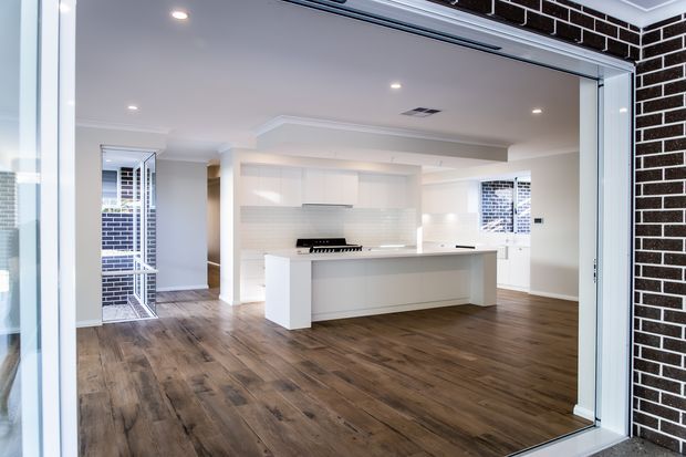 Bright contemporary open-plan kitchen viewed through wide glazed doors. The room features a large white island with integrated cooktop, sleek white cabinetry, a pale tiled splashback, recessed ceiling lights, and warm brown timber-look flooring. Dark brickwork is visible on the exterior surround, while additional glazing and a partially visible adjacent area appear beyond the kitchen.