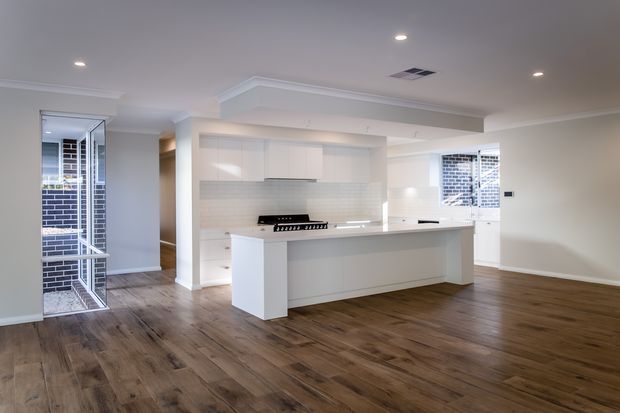 Bright open-plan kitchen with a large white island, white flat-panel cabinetry, pale tiled splashback and integrated stainless-steel appliances. The room has dark timber flooring, white walls with simple cornice detailing, recessed ceiling lights and extensive windows with security screens. The space appears newly renovated and minimally furnished.