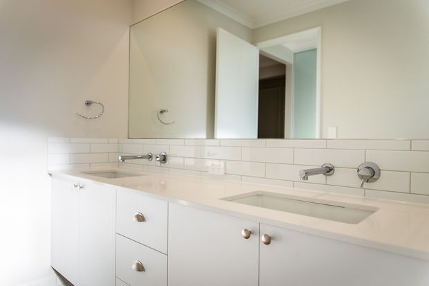 Bright, clean bathroom vanity with a long white built-in cabinet, pale stone benchtop, integrated rectangular basin, chrome wall-mounted tapware, white subway-tile splashback, and a large mirrored wall cabinet. An open door leads to an adjacent space.