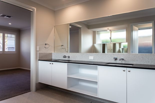 Bright, functional bathroom vanity with a long white built-in cabinet, speckled light stone benchtop, wall-to-wall mirror, twin basins and taps, and open shelving beneath the counter. A carpeted bedroom or hallway is visible through the doorway, while frosted and translucent windows provide natural light.