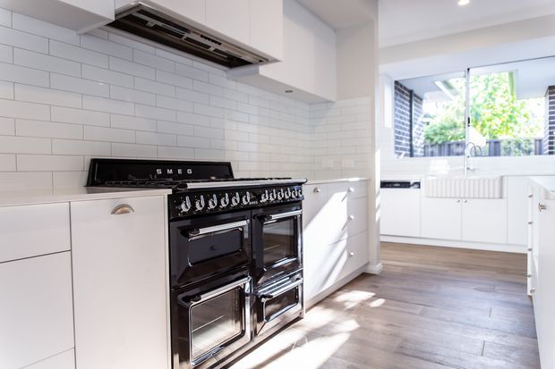 Bright contemporary white kitchen featuring a large black Smeg range cooker, white shaker-style cabinetry, white subway-tile splashback, stainless-steel extractor hood, and medium-toned timber flooring. A glazed opening connects visually to a bright utility or secondary kitchen area.