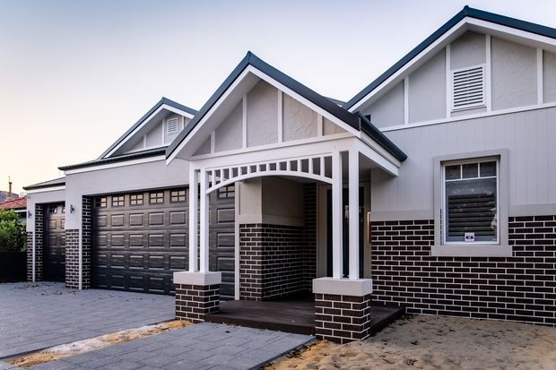 Street-facing view of a newly built single-storey residential house with multiple gabled roof sections, light rendered and weatherboard-style walls, dark brick lower walls, a double garage, and a covered front entry porch with decorative white posts and arch detailing. The driveway and front landscaping are partially unfinished.