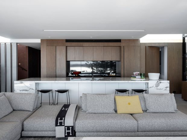 Open-plan contemporary living room with a large grey sectional sofa in the foreground facing a white marble kitchen island and integrated kitchen joinery. The kitchen features warm timber cabinetry, a dark reflective splashback or appliance wall, white cabinetry, black bar stools, and recessed linear ceiling lighting. A hallway or doorway is visible to the left and additional timber-clad joinery appears on the right.