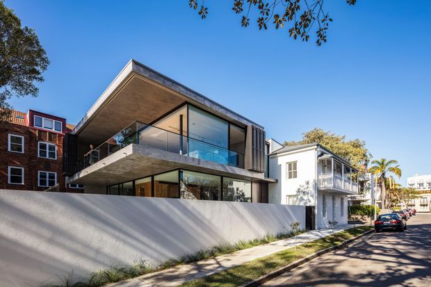 Street-level exterior view of a contemporary multi-level house, featuring a projecting concrete upper floor and balcony, extensive floor-to-ceiling glazing, dark vertical cladding, and a low rendered boundary wall. Older neighbouring homes and an urban residential streetscape are visible around it.