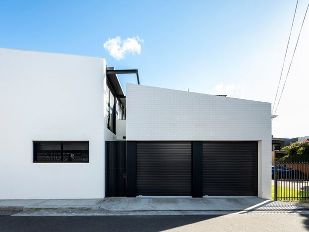 Street-facing view of a contemporary multi-level residence with a crisp white rendered facade, a projecting upper volume clad in small white tiles, black-framed glazing, and two wide dark roller garage doors. The building presents a clean, rectilinear composition with a narrow balcony or recessed entry zone and a concrete driveway frontage.