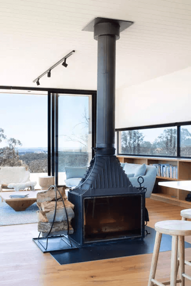 Bright, open-plan mid-century-inspired living room centred around a freestanding black metal wood-burning fireplace with a tall cylindrical flue. The room features extensive black-framed glazing, warm timber flooring, built-in timber shelving, pale upholstered seating, and views of a natural landscape beyond.