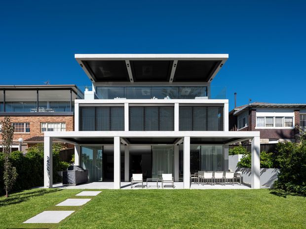 Front exterior view of a contemporary multi-level residence with a crisp white rendered facade, extensive glazing, dark horizontal window screens, and a prominent flat roof canopy. The ground level opens onto a covered outdoor entertaining area with outdoor furniture, leading to a broad manicured lawn and stepping-stone path. Neighbouring suburban homes are visible on either side beneath a clear blue sky.