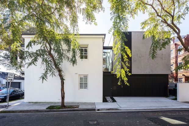 Street-facing view of a compact contemporary urban residence. The facade combines a smooth white rendered two-storey volume with a dark ground-level garage or entry zone and a projecting upper box finished in grey textured cladding. Large areas of glazing connect the volumes, while a mature street tree partially screens the building. Adjacent masonry buildings and a one-way street sign indicate a dense urban setting.
