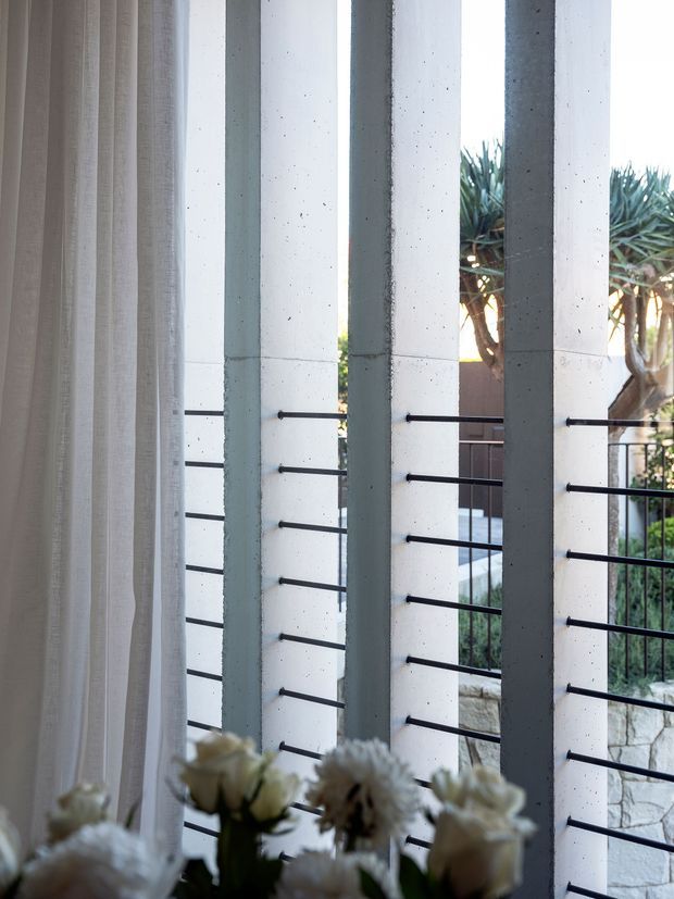 Interior view through floor-to-ceiling glazing with sheer curtains at left, looking toward a covered outdoor area framed by tall pale concrete columns and slim black horizontal metal balustrade rails. A landscaped garden and stone retaining wall are visible outside. The image emphasizes the architectural screen, natural light, and connection between interior and exterior.