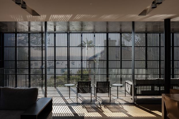 A contemporary interior lounge or living space with extensive floor-to-ceiling glazing screened by dense vertical black slats. Strong daylight casts dramatic striped shadows across the pale floor, ceiling, and furniture. The room includes a dark sofa, low tables, lounge chairs, and a dark built-in bench or seating area, with a small outdoor garden visible beyond the glazing.