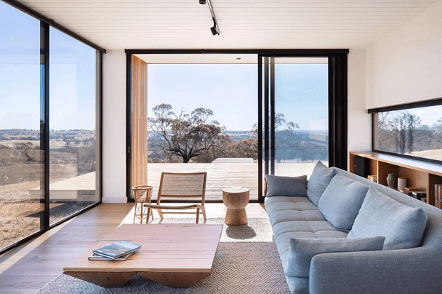 A bright contemporary living room with expansive floor-to-ceiling glazing overlooking a dry, hilly landscape. The room features a large pale blue-grey sectional sofa, low timber coffee table, woven rug, woven lounge chair, and built-in timber shelving beneath a horizontal window. Black-framed sliding glass doors open to a broad outdoor terrace, while a linear track light runs across the white paneled ceiling.