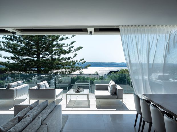 A bright, contemporary open-plan living and dining area with floor-to-ceiling glazing opening onto a balcony or terrace. Pale upholstered sofas and armchairs surround a low glass coffee table, while a dining table with dark chairs sits at the right. Sheer white curtains, a polished light floor, and extensive glazing frame views of the ocean and coastal landscape, creating an airy indoor-outdoor setting.