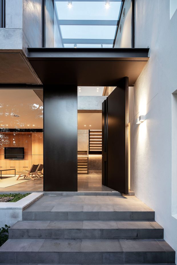 A modern residential entry viewed from outside, with broad dark grey stone steps leading to a tall black pivot door and adjacent full-height glazing. The entrance is framed by white rendered walls, a black canopy, and a glazed skylight above. Through the open doorway and glass, a warm-toned interior with timber wall finishes, illuminated shelving or stair elements, and lounge seating is visible. The composition has strong vertical lines, contrasting dark metalwork, pale walls, and warm interior lighting.