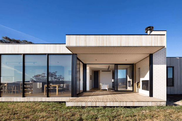Exterior view of a contemporary single-storey home with a strong horizontal composition, pale vertical cladding, dark-framed floor-to-ceiling glazing, a sheltered timber deck, and a recessed outdoor living area with a built-in fireplace. The image shows a clean-lined flat-roof form against a clear blue sky.