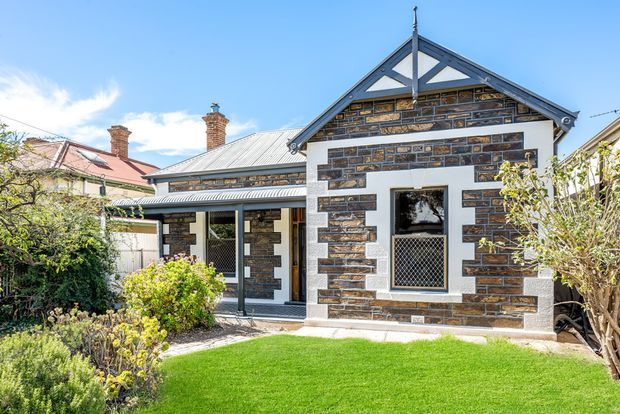 Front exterior of a single-storey heritage-style brick cottage with a gabled roof, decorative timber gable detailing, contrasting light mortar and rendered trim, a covered entry porch, and a landscaped front garden. The facade features dark patterned brickwork, a corrugated metal roof, and traditional chimneys.