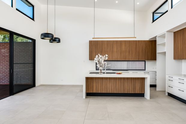 Bright, double-height contemporary kitchen with a large central island, white walls, pale tiled flooring, and extensive warm timber cabinetry. Black-framed glazing and a glazed external door open to a brick-lined outdoor area. The kitchen includes white benchtops, integrated shelving, a ribbed splashback, black pendant lights, and a linear timber pendant above the island.