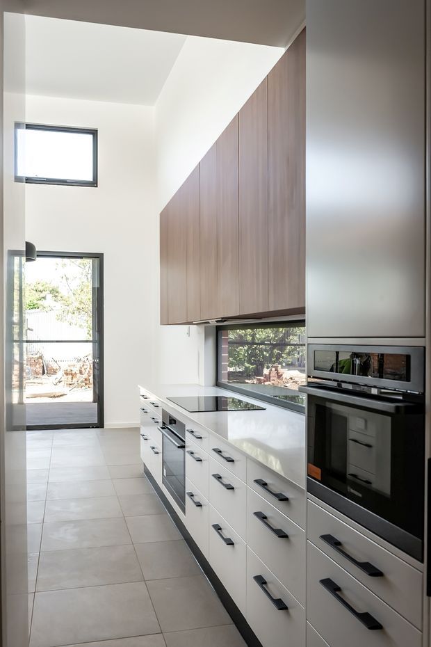 A bright, narrow modern kitchen with a tall ceiling and strong natural light. The space features extensive white drawer cabinetry with black horizontal handles, a dark cooktop and integrated appliances, a pale stone or solid-surface benchtop, and a full-height vertical timber-look cabinetry wall beneath the sloping ceiling. Large black-framed windows and a glazed door bring in views of the outdoors. Large light-grey floor tiles complete the clean, contemporary fit-out.