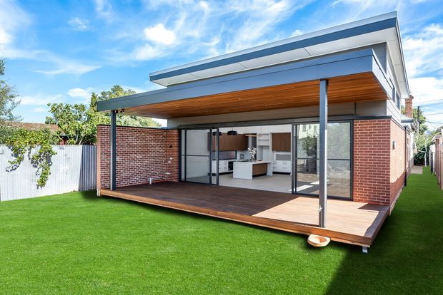 Contemporary single-storey extension viewed from the rear garden, featuring a flat roof with broad eaves, red brick walls, extensive black-framed glazing, and a covered timber-lined outdoor terrace. The open doors reveal a bright kitchen and living area, while the landscaped lawn surrounds the deck.