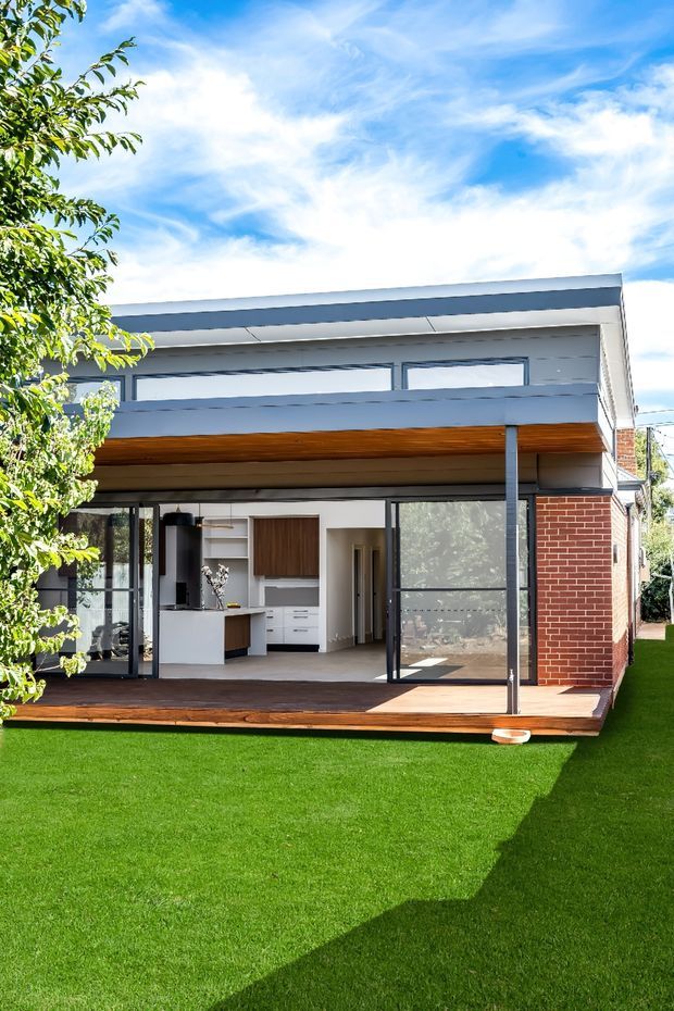 Bright exterior view of a contemporary two-storey residential extension with a flat roof, red brick side wall, white rendered upper facade, extensive dark-framed glazing, and a warm timber soffit over a timber deck. The open ground level reveals a compact kitchen and interior hallway, while a manicured lawn occupies the foreground.