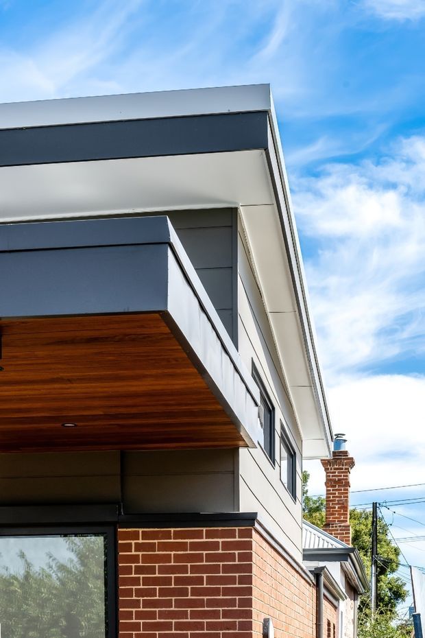 Close-up exterior architectural view of a contemporary multi-level home, showing a crisp flat roofline, projecting upper-floor volume, dark grey cladding, white soffits and trims, a warm timber-lined cantilevered canopy, and a red brick lower facade. A chimney and portions of the neighbouring streetscape are visible at the side under a bright blue sky.