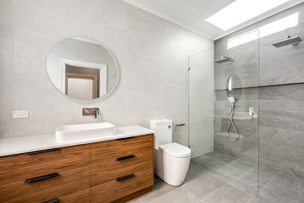 Contemporary bathroom with a floating timber vanity, white rectangular vessel basin, wall-mounted tapware, round mirror, toilet, and a large frameless glass walk-in shower. The shower and floor are finished in grey stone-look tiles, with pale grey wall tiles, a skylight, and clean minimalist detailing.