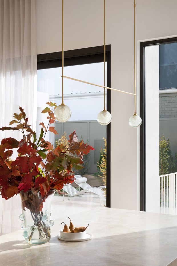 Bright, refined kitchen interior featuring a pale stone island or benchtop, tall black-framed windows, sheer curtains, and three suspended globe pendant lights with brass-toned slender fittings. A large autumnal floral arrangement and fruit bowl sit on the island. The glazing looks onto a private courtyard with outdoor loungers and a rendered or brick boundary wall.