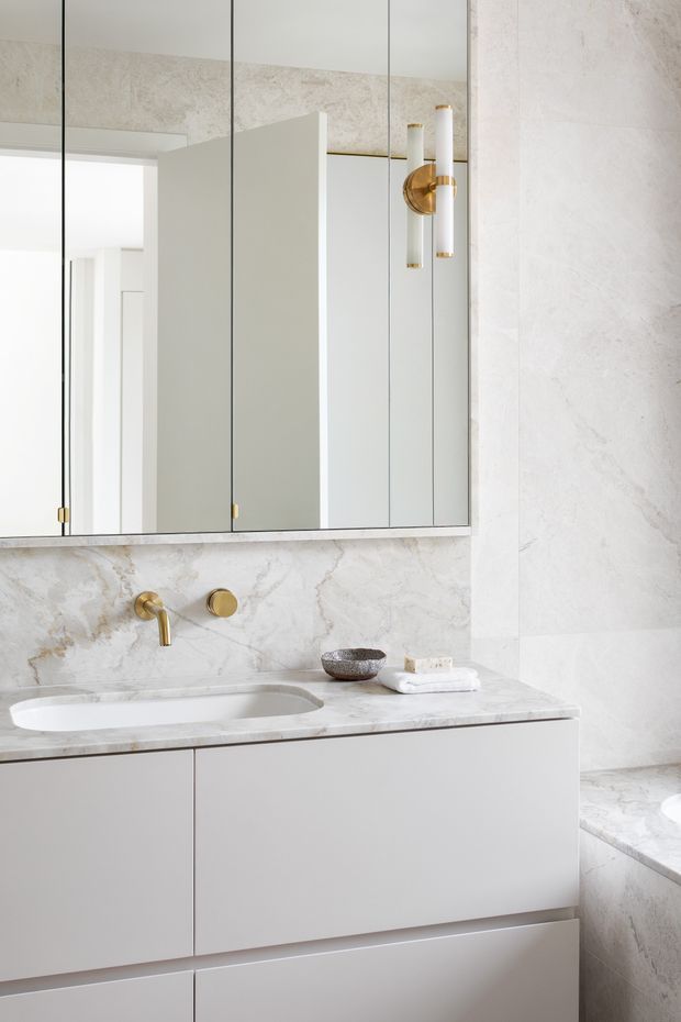 Refined contemporary bathroom vanity with a pale marble or stone wall finish, matching stone benchtop and integrated basin, minimalist light-grey cabinetry, a large mirrored cabinet, and warm brass wall-mounted tapware and lighting. A partial bathtub is visible at right.