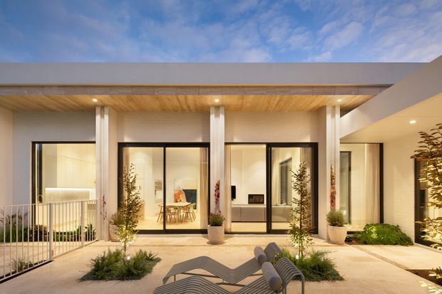 Contemporary single-storey residence viewed from a landscaped courtyard or patio at dusk. The facade features broad floor-to-ceiling glazing with dark frames, slender white structural columns, a warm timber-lined soffit, pale rendered walls, and a sheltered outdoor seating area with integrated planting.