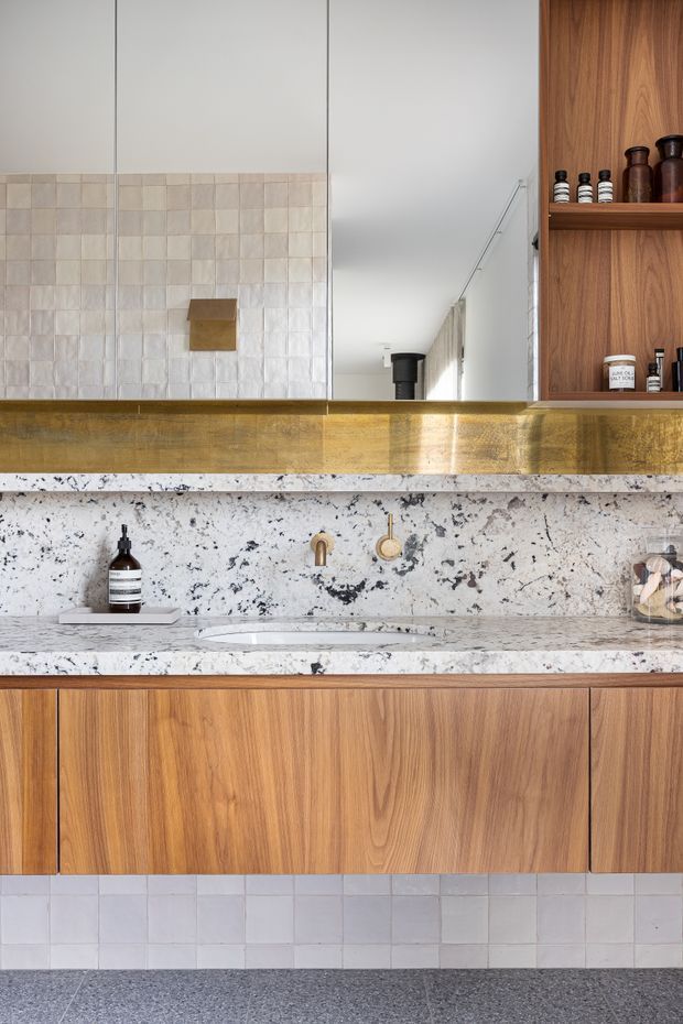 A refined bathroom vanity featuring warm timber cabinetry, a white-and-grey veined stone countertop and backsplash, integrated basin, brushed brass tapware, a brass-framed mirror, and open timber shelving with toiletries. The wall behind the mirror and vanity is finished with pale square tiles, while the floor is a dark speckled surface.