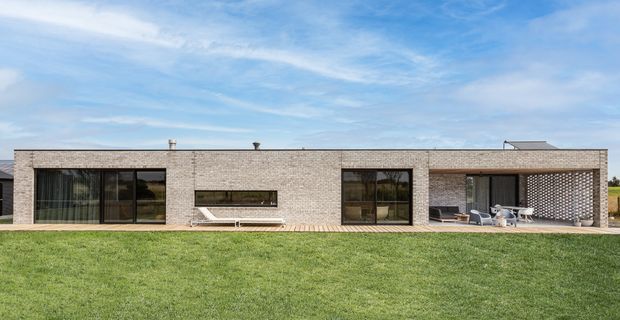 A low, single-storey contemporary building viewed from the lawn, with a long pale brick facade, broad dark-framed glazed openings, a recessed outdoor terrace, and a perforated brick privacy screen at the right. The flat roof and restrained rectangular composition create a minimalist, modern exterior.
