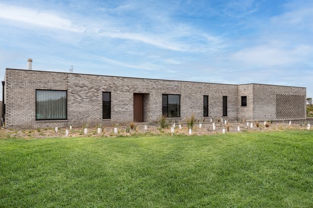 Single-storey contemporary brick building with a long, low rectangular form, flat roofline, narrow dark-framed windows, a recessed entry, and a perforated brick screen at the right end. The building sits behind a landscaped foreground lawn under a blue sky.