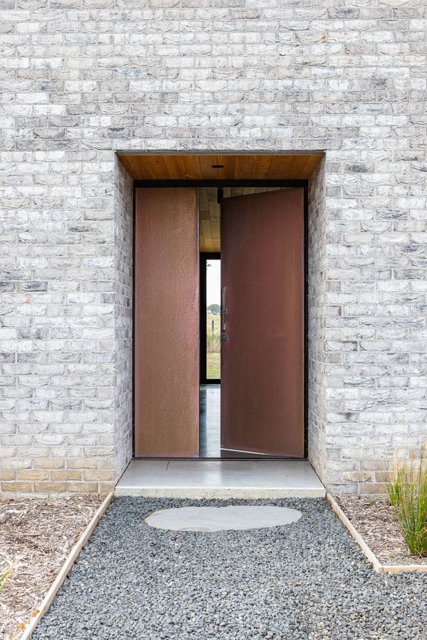 Front exterior entry of a contemporary building, featuring a pale white-grey brick facade, a deep recessed doorway with large rust-brown pivot doors, a timber-lined soffit, and a gravel path with a circular concrete stepping stone.