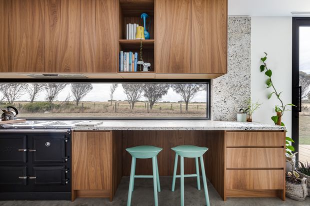 A contemporary kitchen with extensive warm timber cabinetry, an open shelf niche, a long horizontal picture window, pale stone benchtops, and a black freestanding range cooker. Two mint-green stools sit at a compact peninsula, with a rural landscape visible beyond the glazing. A textured pale stone feature wall and indoor plants add natural character.