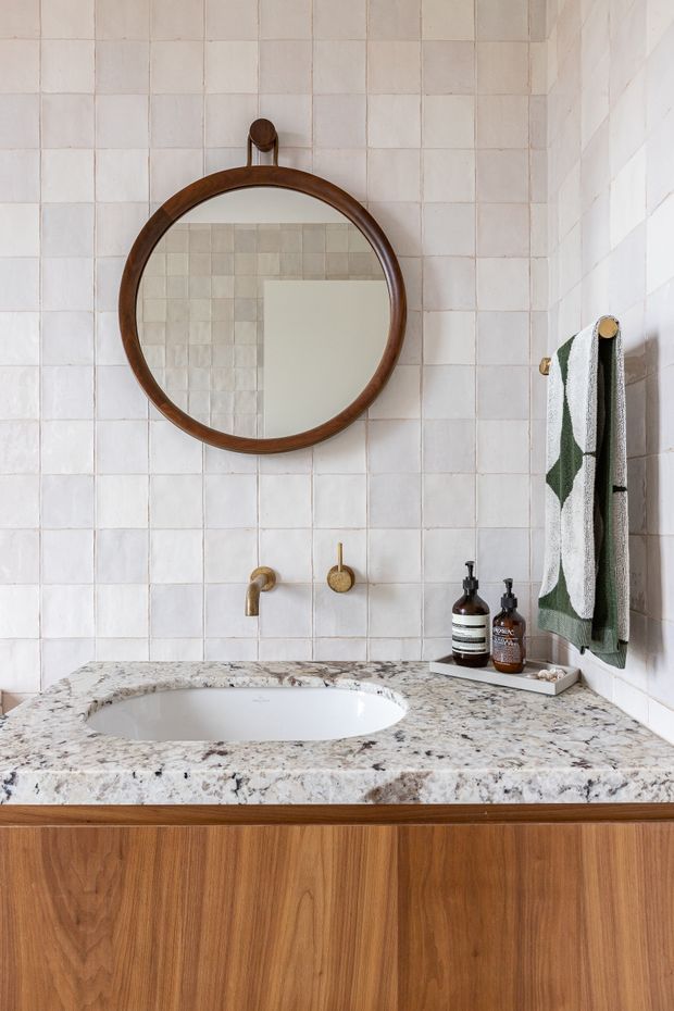 Close-up view of a refined bathroom vanity. The space features pale square tiled walls, a round dark timber-framed mirror, a heavily veined natural stone countertop with an integrated white basin, brushed brass wall-mounted tapware, and warm timber cabinetry below. A patterned towel and toiletries add restrained decorative detail.