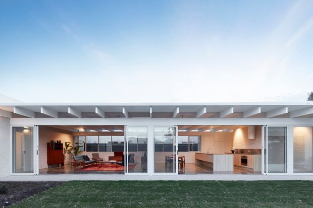 Exterior view of a low, single-storey contemporary house with extensive floor-to-ceiling glazing and a broad flat roof with exposed projecting beams. The white facade opens onto a landscaped lawn, revealing a warm open-plan living area and a modern kitchen inside.
