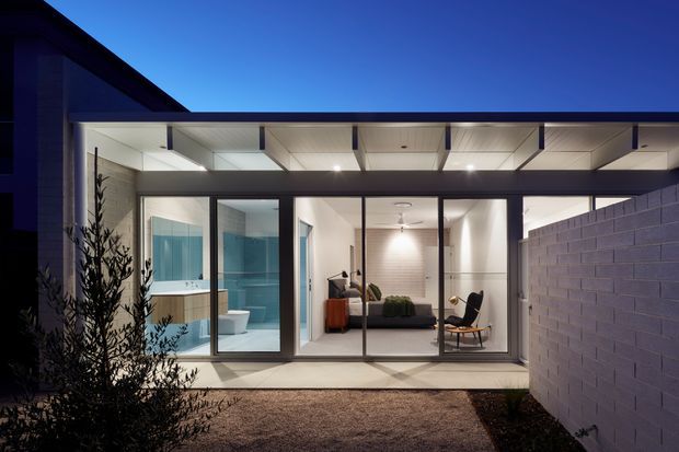 Twilight exterior view of a contemporary single-storey residence, focused on a glazed rear or side extension. Full-height glass doors and windows reveal a bright bathroom and adjacent living or sitting area, while a projecting flat roof with exposed structural framing forms a covered terrace. Dark cladding contrasts with a pale rendered or painted brick boundary wall and simple landscaped courtyard.