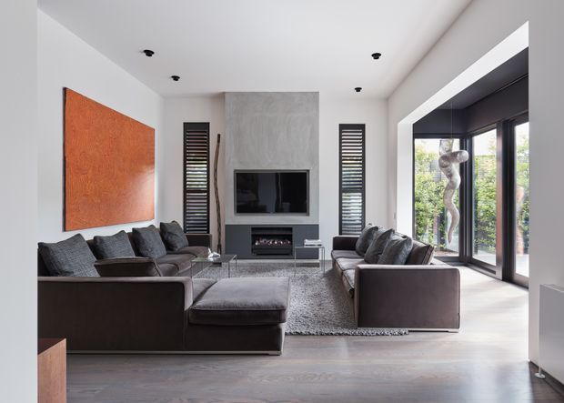 A refined contemporary living room with a large dark sectional sofa and matching armchairs arranged around a glass coffee table. The room features a wall-mounted television set into a textured grey feature wall above a linear fireplace, dark louvred window panels, expansive black-framed glazing, grey timber-look flooring, and a large warm terracotta artwork. The space is bright, spacious, and carefully balanced with neutral grey and charcoal furnishings.